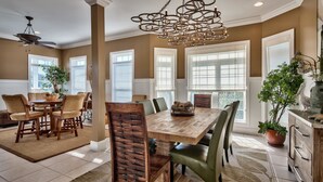 A spacious dining area with a large wooden table and chairs, surrounded by large windows and a decorative chandelier.