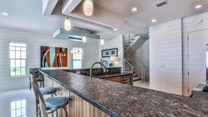 A modern kitchen with a long island, bar stools, pendant lighting, and a staircase leading to an upper level. - Bord du Mer 6 Bedroom Holiday Home by Five Star Properties (Miramar Beach)