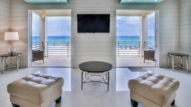 A modern living room with a large TV, two leather ottomans, a round coffee table, and a view of the beach through large windows. - Bord du Mer 6 Bedroom Holiday Home by Five Star Properties (Miramar Beach)