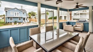 A covered outdoor patio with a glass table and beige chairs, overlooking a residential area with a blue house. - Bottoms Up 7 Bedroom Holiday Home by Five Star Properties (Destin)