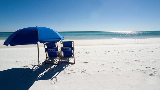 Ein Strand mit zwei blauen Liegestühlen unter einem blauen Sonnenschirm und Fußabdrücken im Sand.