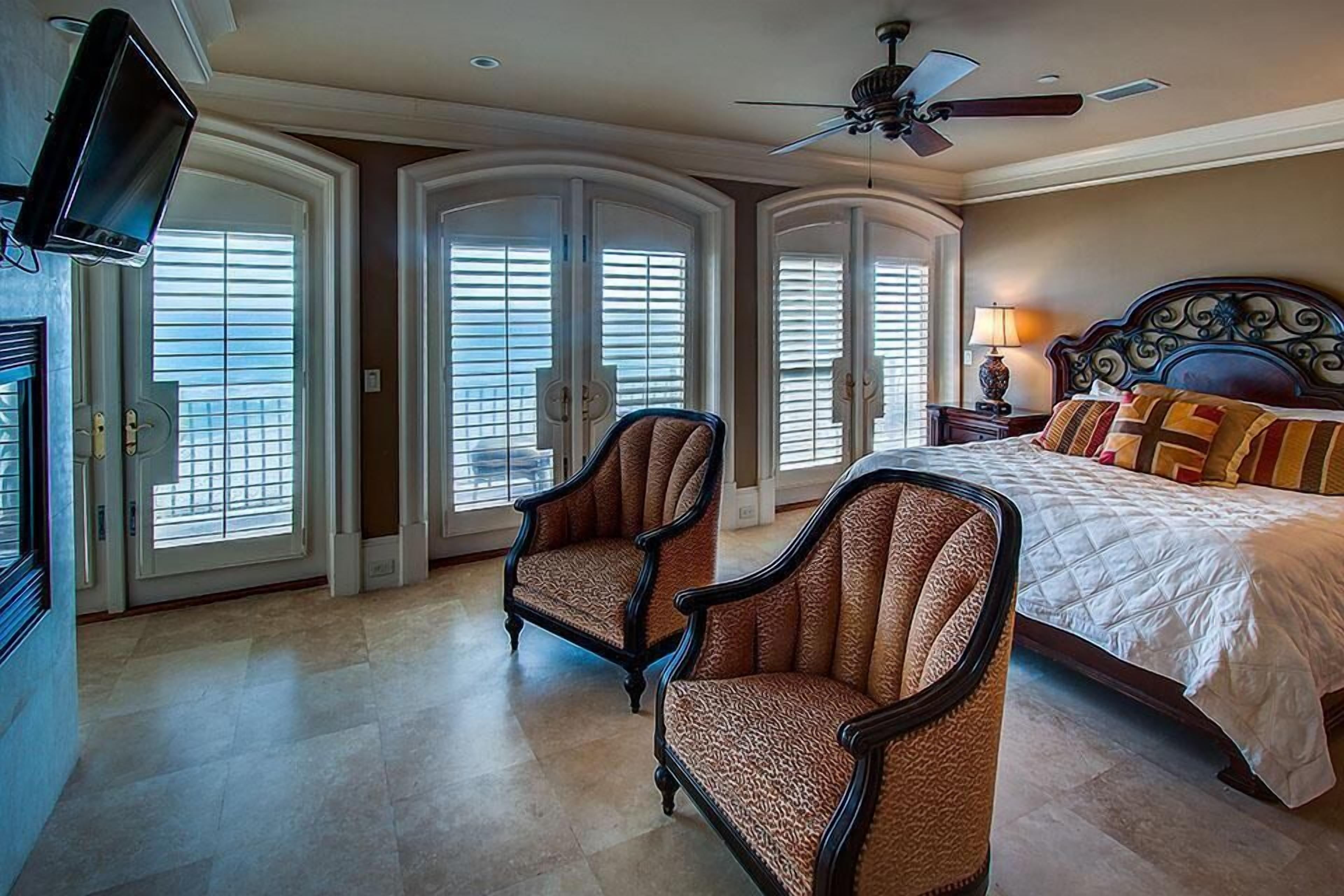 A spacious bedroom with a large bed, two armchairs, a ceiling fan, and a TV mounted on the wall.