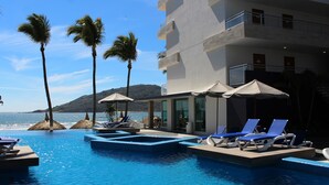 Zona de piscina con sillas de descanso y sombrillas, con vista a una playa y el océano.