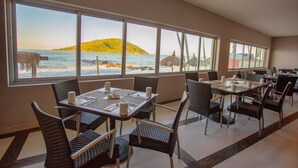 Comedor con amplias ventanas que dan a una playa, con sillas y mesas de mimbre dispuestas para comer.