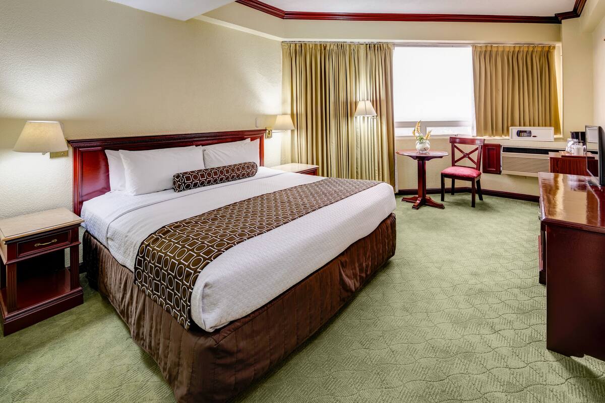 Habitación de hotel con una cama grande, un escritorio, una silla, una lámpara y un ventanal con cortinas.