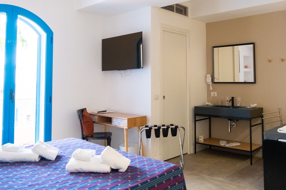 A hotel room with a bed, a desk, a chair, a TV, a bathroom with a sink and mirror, and a door with a blue frame.