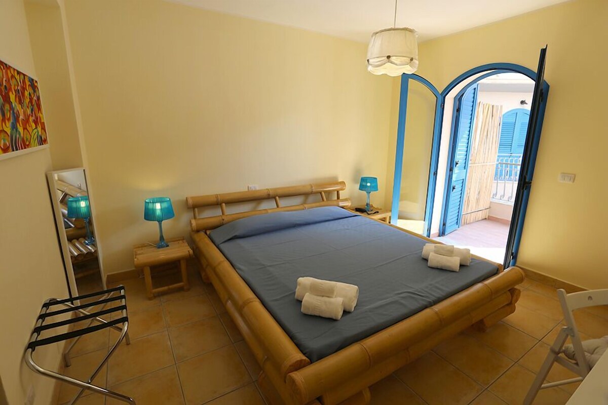 A room with a bed, two bedside lamps, a chair, and a door leading to an outdoor area.