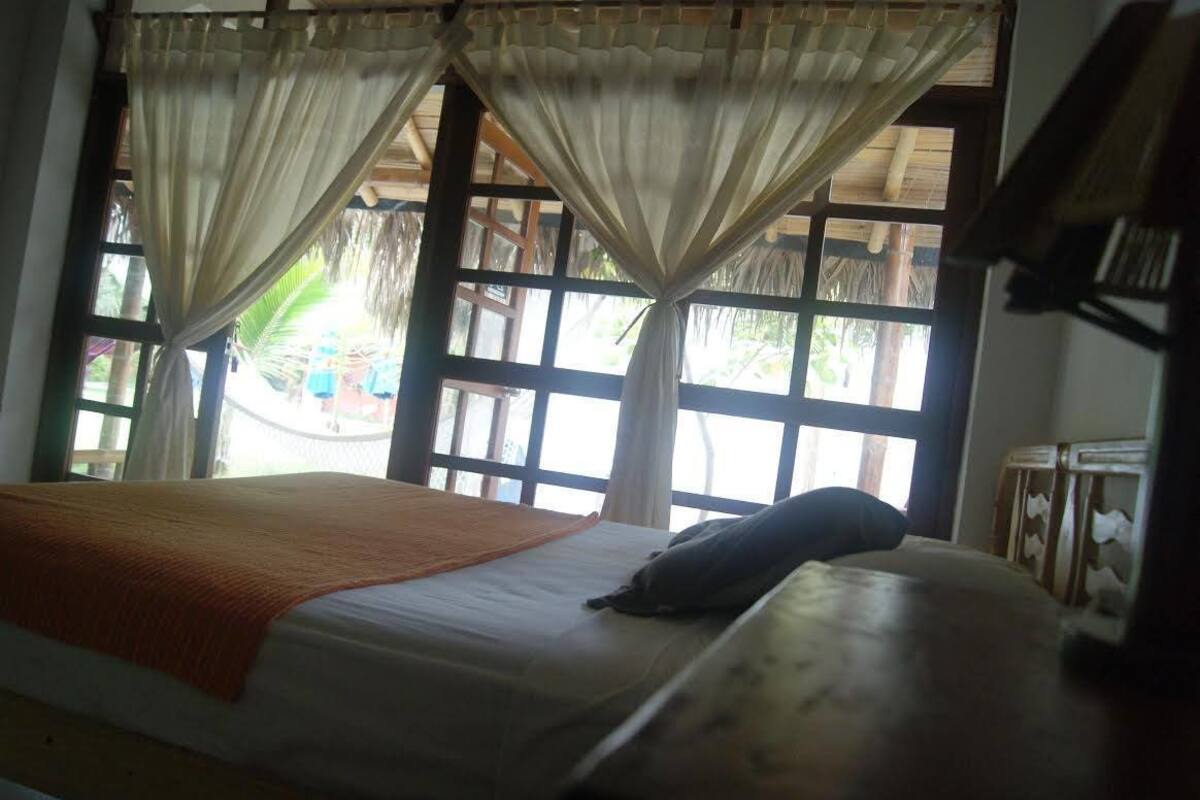 A bed with a white sheet and an orange blanket, a pillow, and a window with curtains.