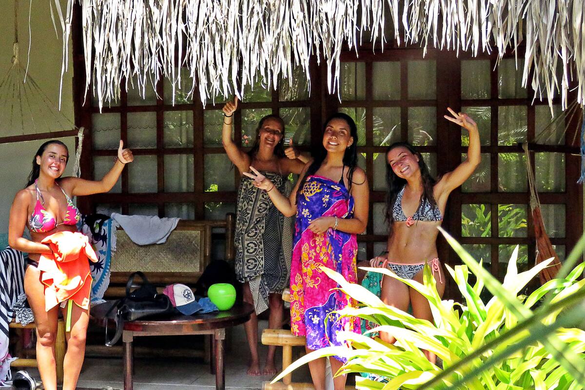 A group of people in a covered outdoor area with a thatched roof, wooden furniture, and a view of greenery through the windows.