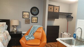 Habitación compacta con cama, una silla, escritorio, cómoda, espejo, reloj y zona de cocina con lavamanos y armarios.