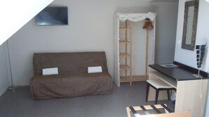 A small, compact room with a brown sofa, a wooden shelf unit, a desk with a chair, and a large mirror. - Sweet Home Appart Hotel Deauville Sud (Saint-Arnoult)