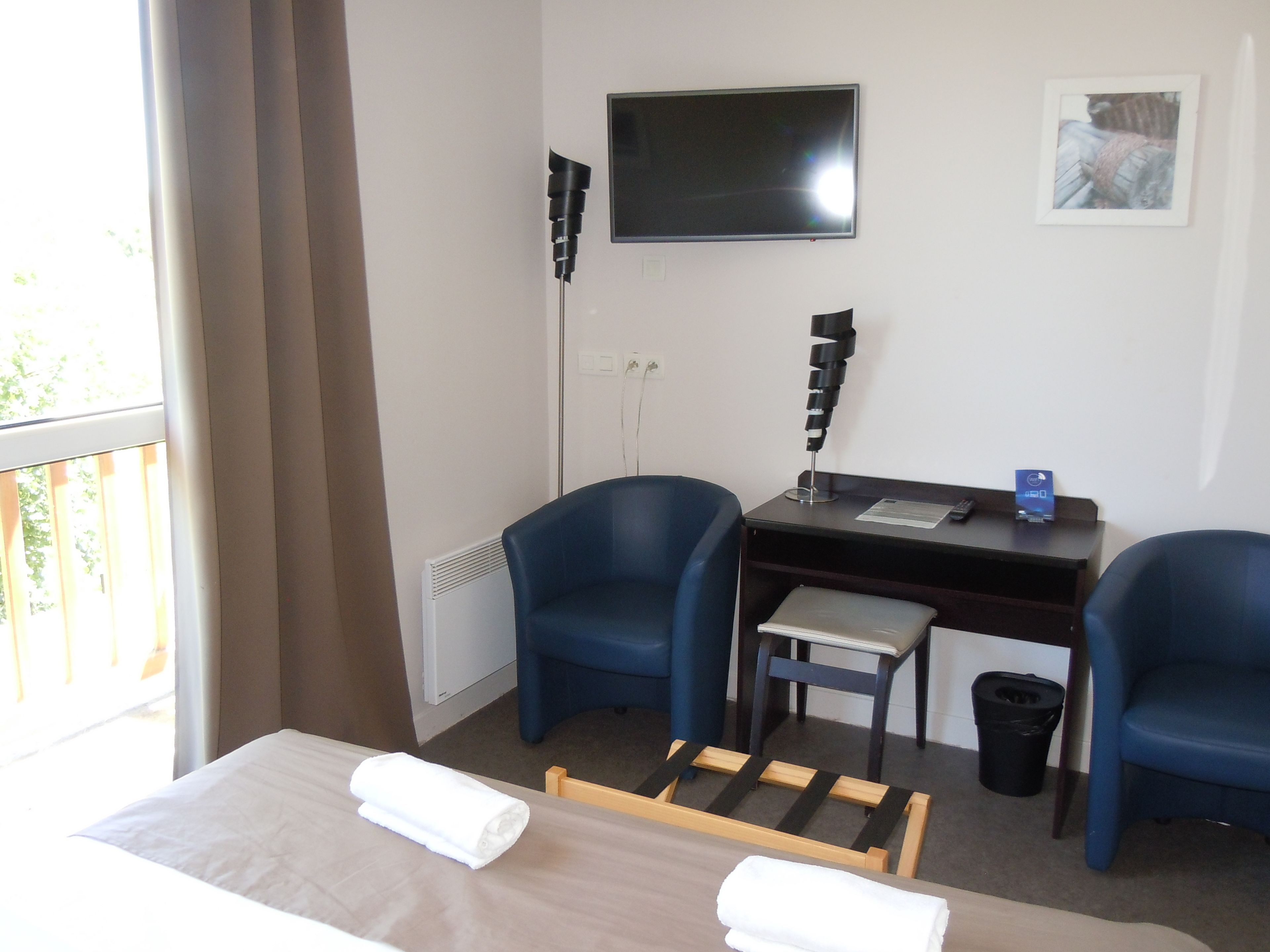 A hotel room with a bed, two blue chairs, a desk with a computer, a TV, and a small window.