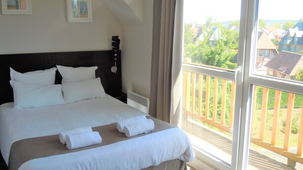A neatly made bed with white linens and towels, a dark headboard, and a large window with a view of houses and greenery. - Sweet Home Appart Hotel Deauville Sud (Saint-Arnoult)