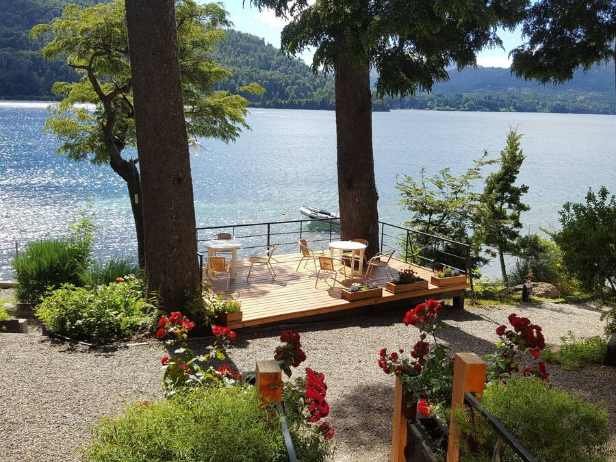 A deck with outdoor seating overlooking a lake, surrounded by trees and a garden with red flowers.