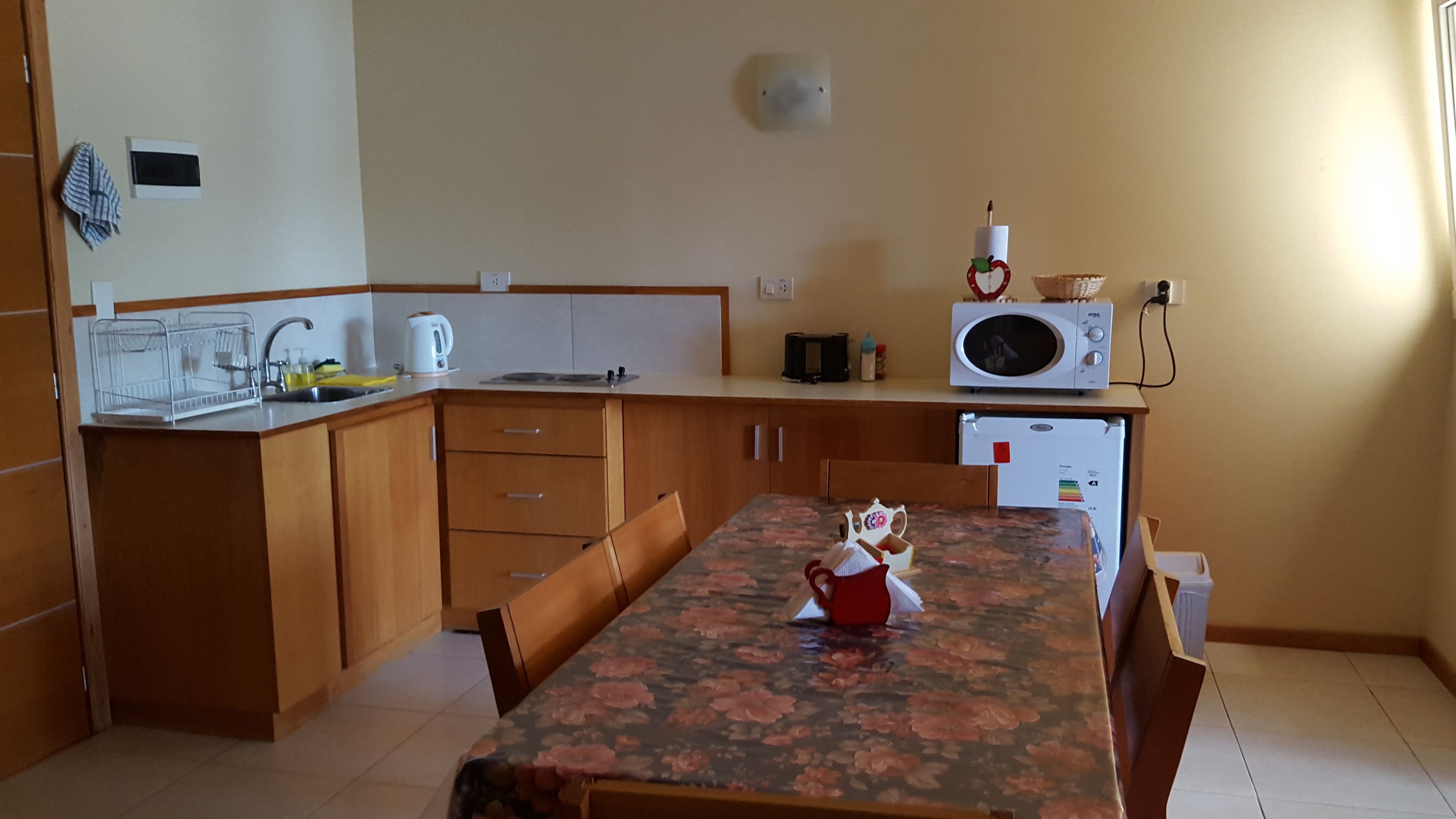A compact kitchen with wooden cabinets, a microwave, a kettle, and a table with a floral tablecloth.
