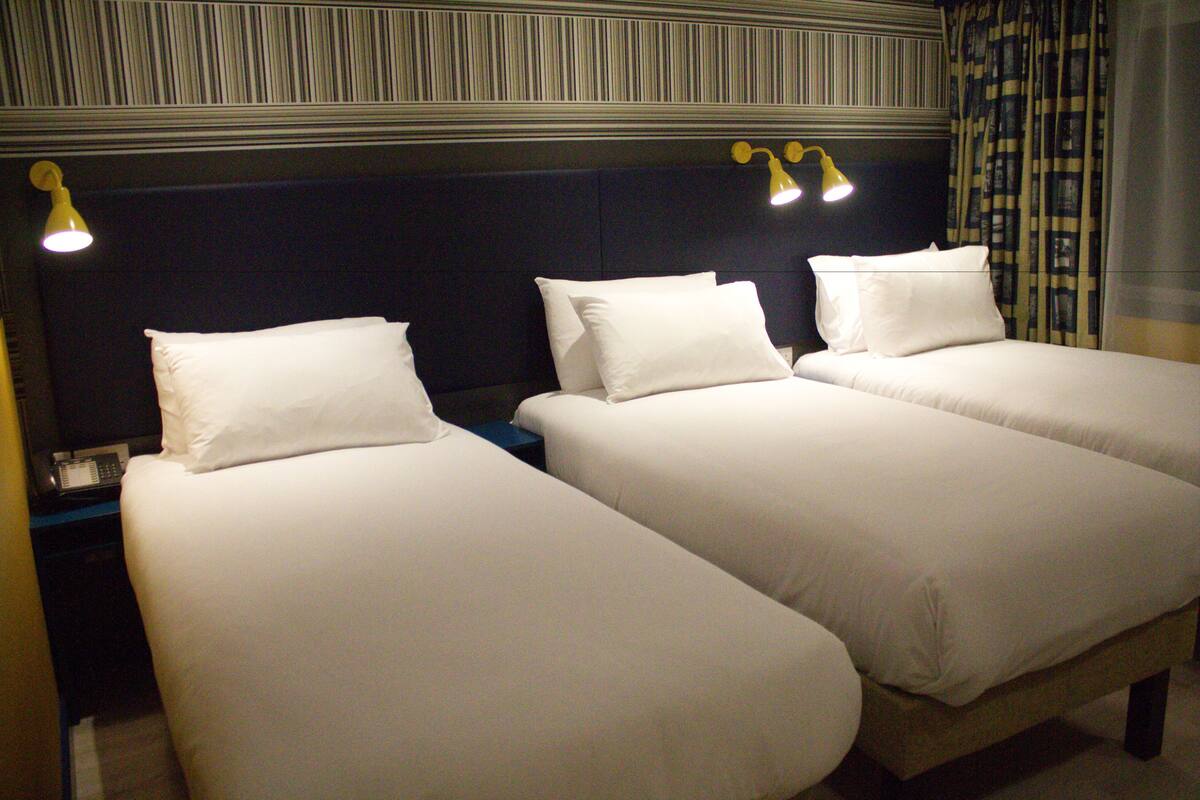 Una habitación de hotel con dos camas individuales, un cabecero oscuro y una mesita de noche con lámpara.