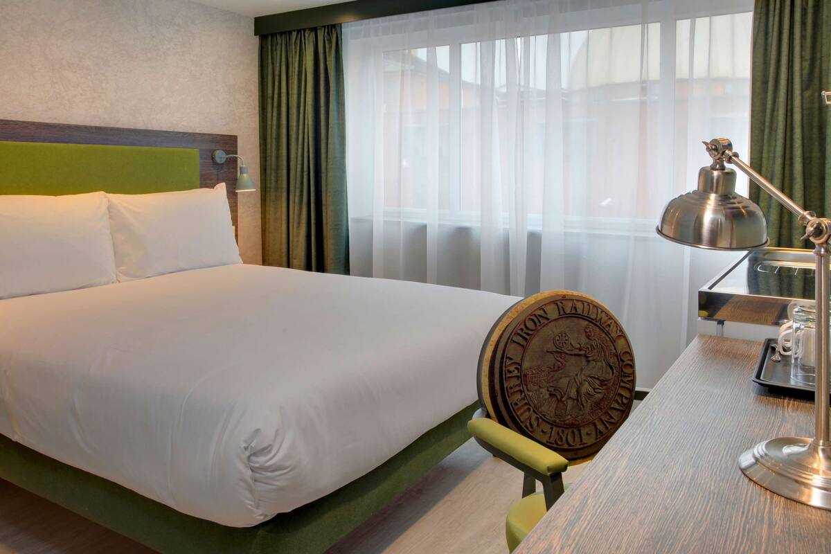 Habitación de hotel con cama, escritorio con lámpara, bandeja con taza y ventana con cortinas translúcidas.