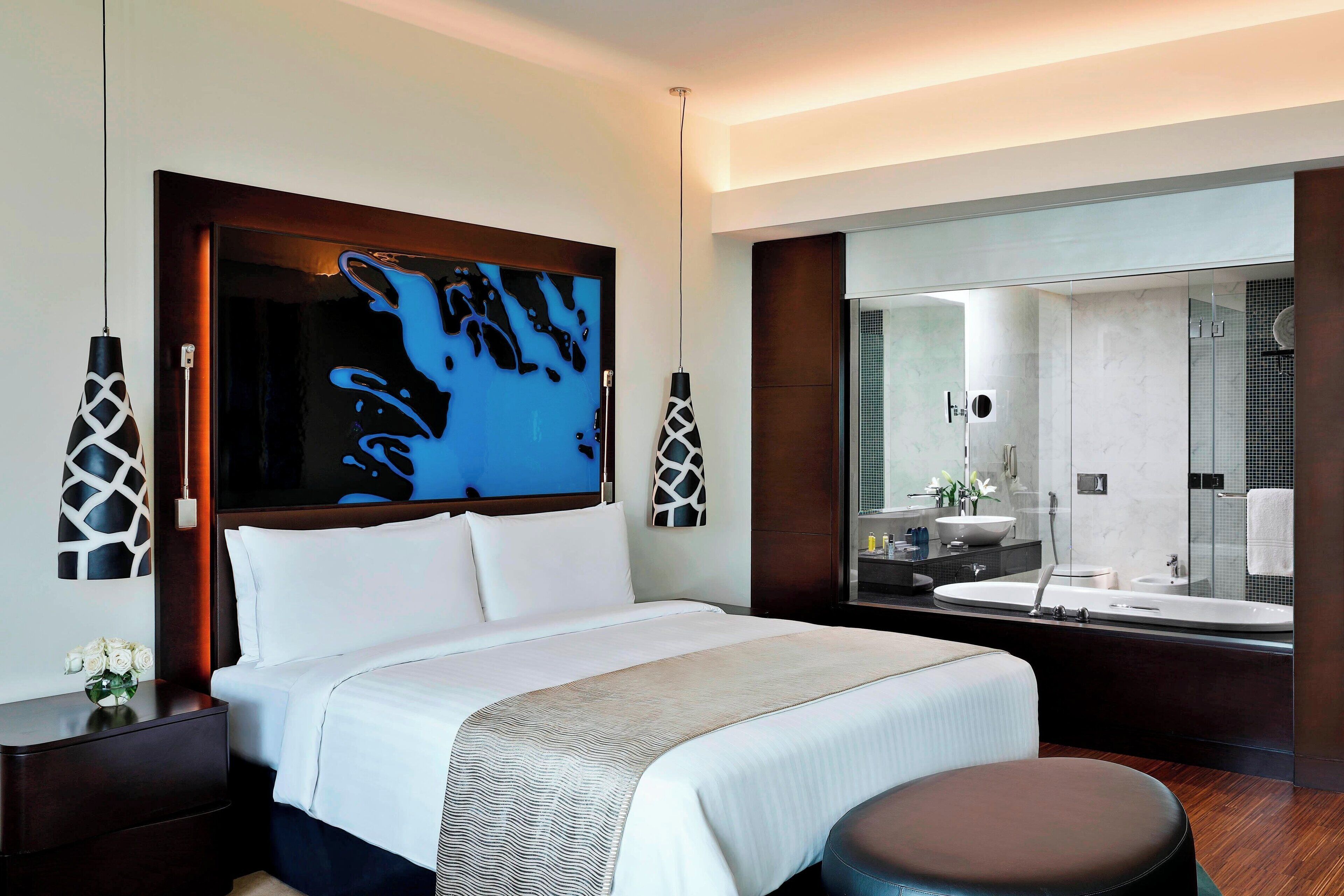 A modern hotel room with a large bed, a sitting area, a bathroom with a large mirror, and artwork on the wall.