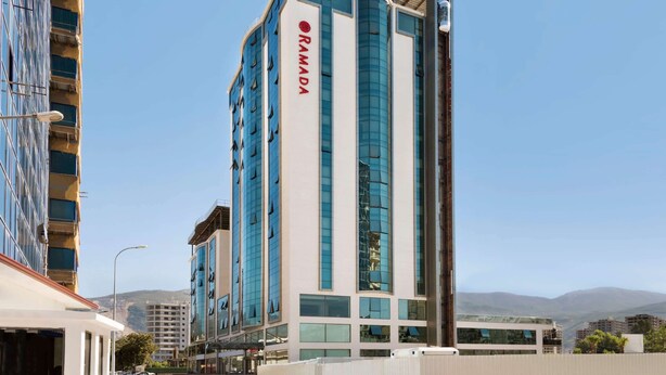 Un hotel moderno de altura con un llamativo letrero 'RAMADA', que presenta una fachada de vidrio y hormigón, y vistas del paisaje urbano circundante.