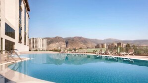 A swimming pool with lounge chairs, a building with large windows, and mountains in the background. - Ramada by Wyndham Iskenderun (Iskenderun)