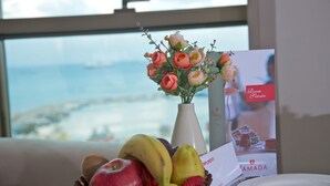 A fruit bowl with bananas, apples, and strawberries, a vase with pink flowers, and a plate on a table with a brochure. - Ramada by Wyndham Iskenderun (Iskenderun)