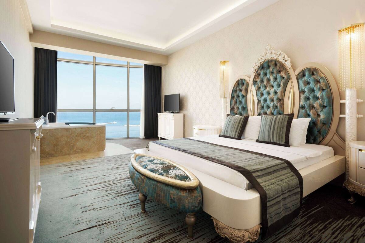 A spacious bedroom with a large bed, a vanity, a television, and a view of the sea.
