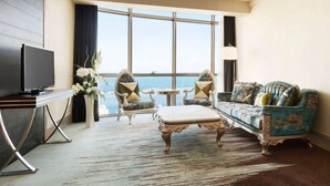 Habitación de hotel con un ventanal grande, televisor, sofá, dos sillones, una mesita y un jarrón decorativo con flores.