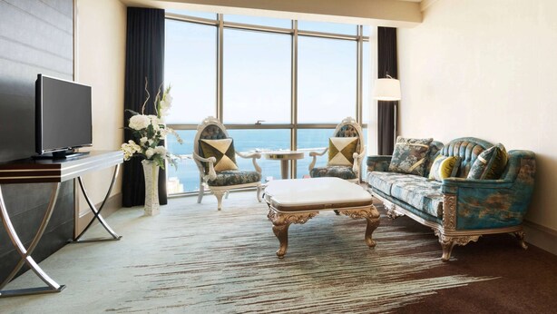 A hotel room with a large window, a television, a sofa, two armchairs, a small table, and a decorative vase with flowers. - Ramada by Wyndham Iskenderun (Iskenderun)