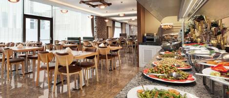 Un comedor moderno con varias mesas y sillas, una barra de buffet con diversos platos, y grandes ventanales que dejan entrar luz natural.