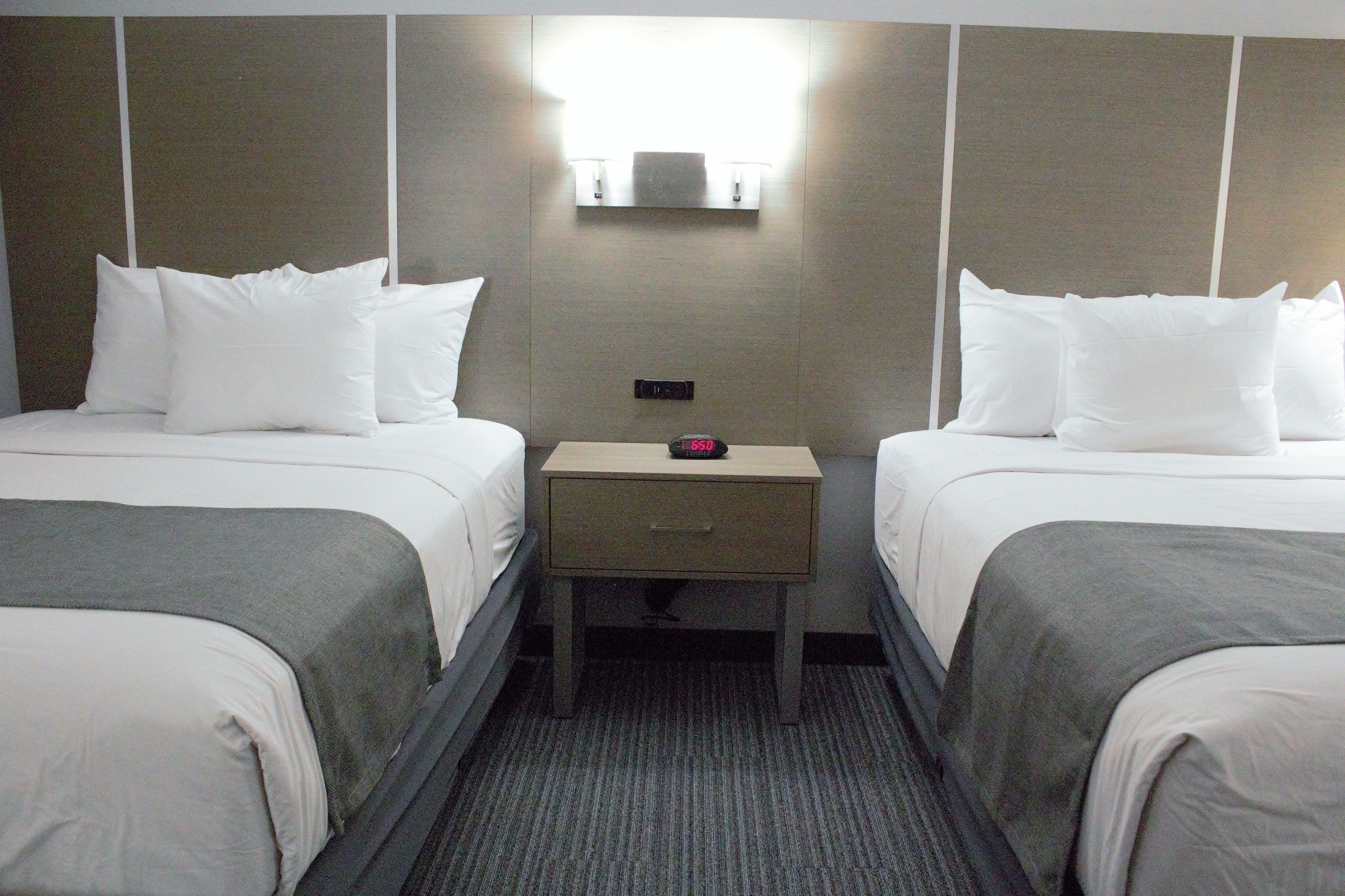A hotel room with two single beds, a nightstand with a clock, and a wall-mounted light fixture.