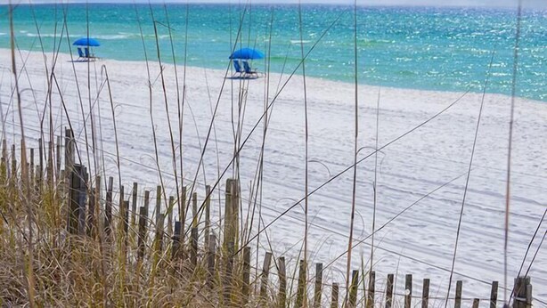 Ein Strand mit weißem Sand und klarem türkisblauem Wasser. Im Vordergrund stehen drei blaue Sonnenschirme und Holzzaun.