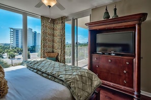Een slaapkamer met een groot bed, een televisie op een houten kast, en een balkon met uitzicht op gebouwen en groen.