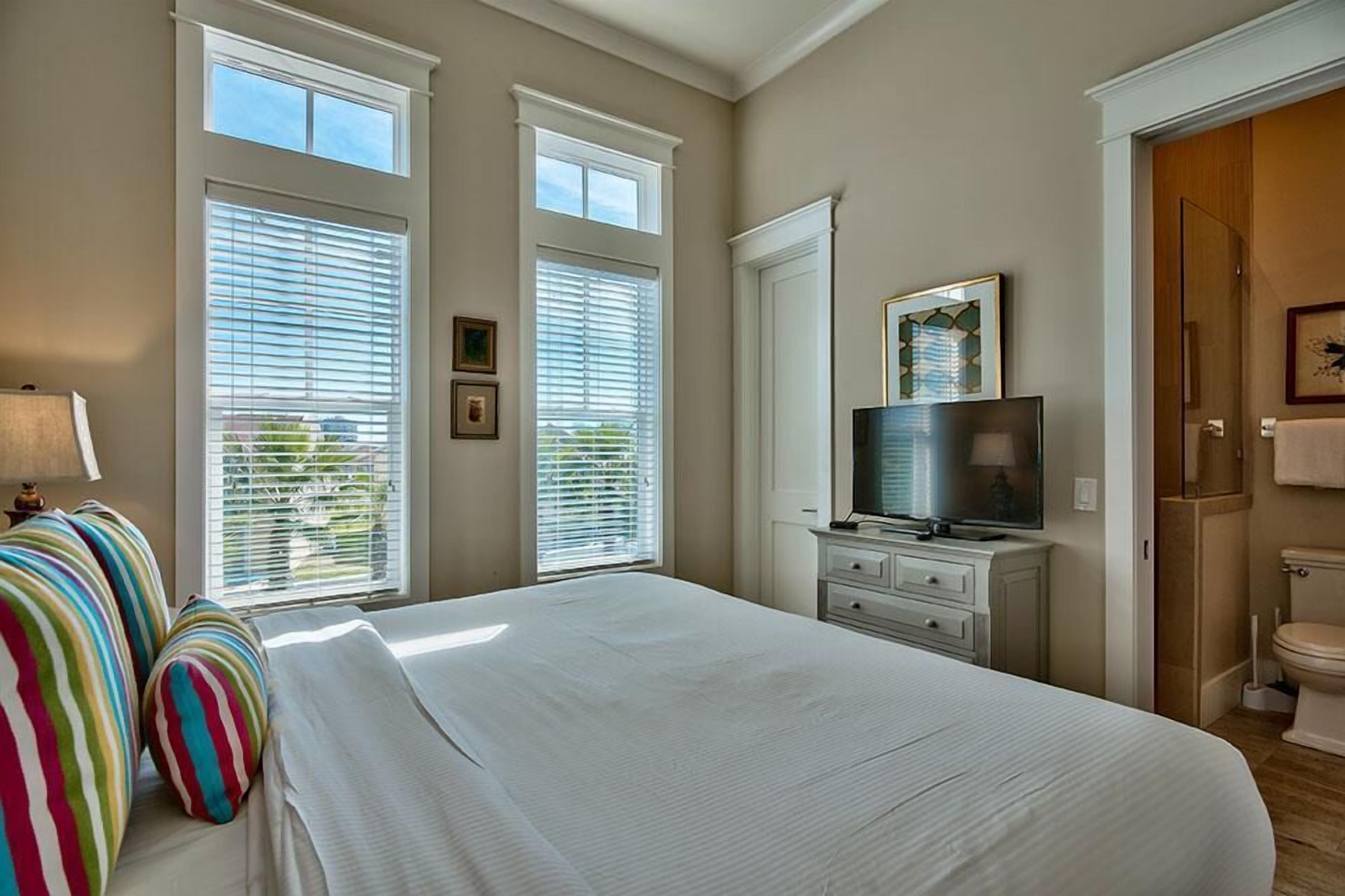 Une chambre à coucher avec un grand lit, deux fenêtres équipées de stores, une télévision posée sur une commode et une salle de bain avec des toilettes.