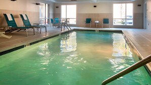 An indoor swimming pool with lounge chairs and tables, large windows, and a tiled floor.