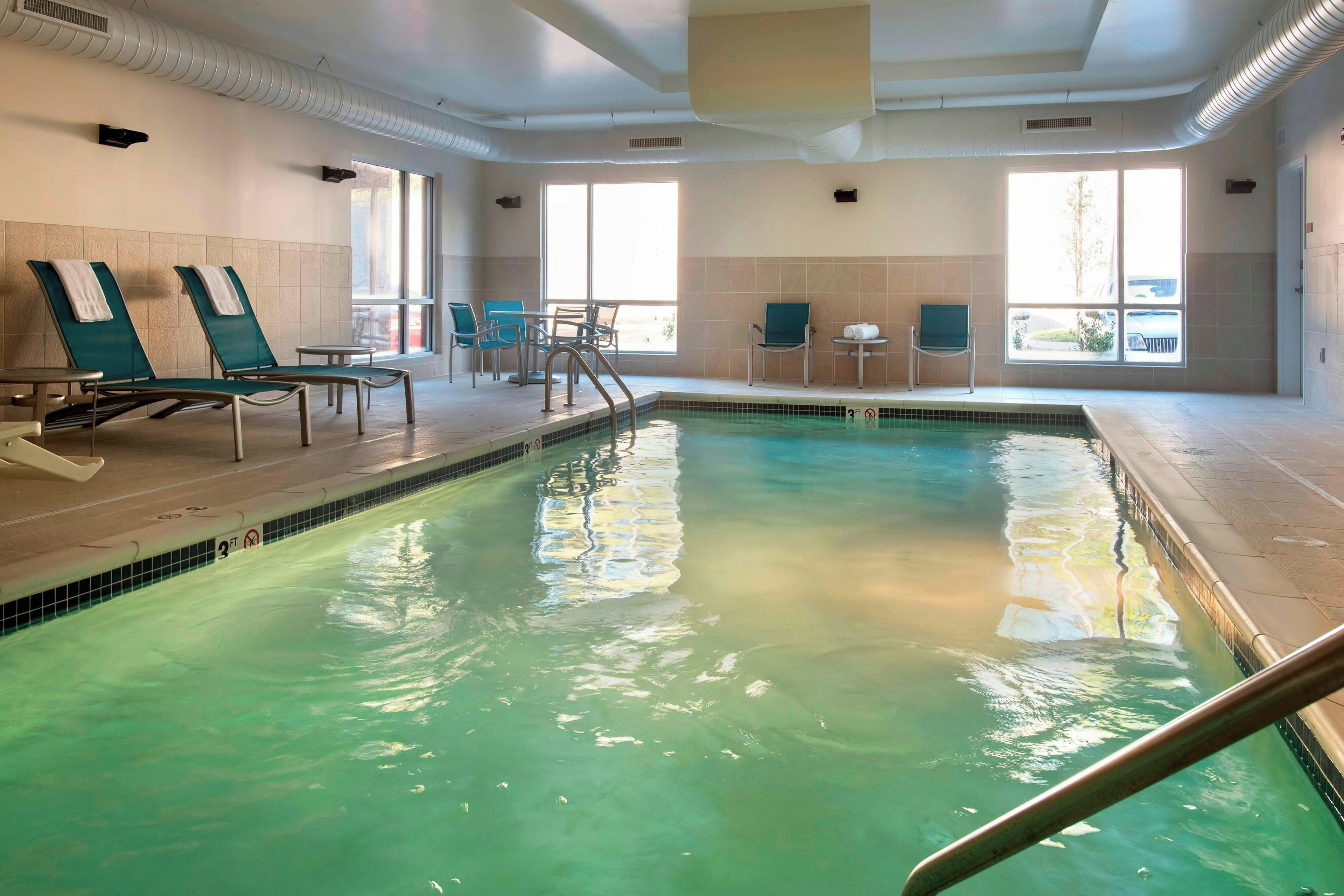 Une piscine intérieure dotée de chaises longues et de tables, d’un sol carrelé et de grandes fenêtres.