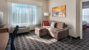 A modern hotel room with a sectional sofa, a desk with a laptop, a standing lamp, and a large window with sheer curtains.