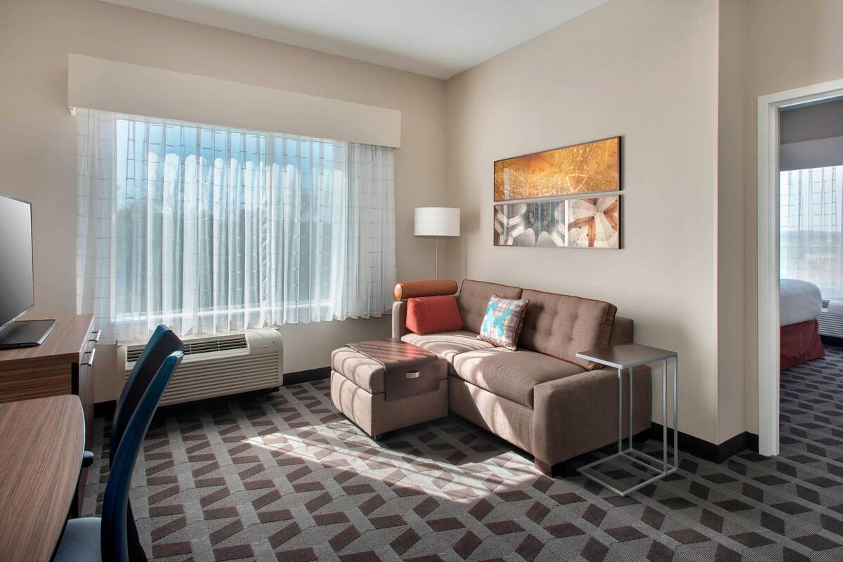 Une chambre d’hôtel moderne, dotée d’un canapé d’angle, d’un bureau avec un ordinateur portable, d’une lampe sur pied et d’une grande fenêtre agrémentée de rideaux légers.