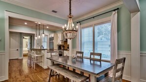 A dining area with a wooden table and benches, a chandelier, and a view of the kitchen and dining room through a window. - Summer Dreams 4 Bedroom Holiday Home by Five Star Properties (Santa Rosa Beach)