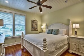 A bedroom with a large bed, a ceiling fan, a nightstand with a lamp, and a window with blinds.