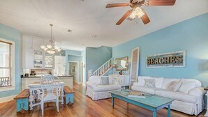 A spacious living room with a wooden floor, white furniture, a ceiling fan, and a kitchen in the background. - GOING COASTAL 3 Bedroom Holiday Home by Five Star Properties (Panama City Beach)