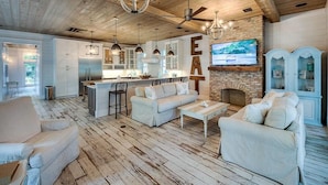Uma sala de estar ampla com piso de madeira, sofás brancos, lareira e cozinha ao fundo.