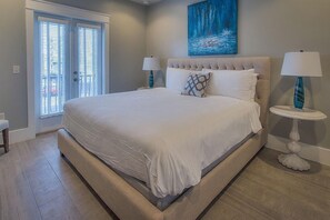 A bedroom with a large bed, two bedside tables with lamps, a painting on the wall, and a sliding glass door leading to an outdoor area.