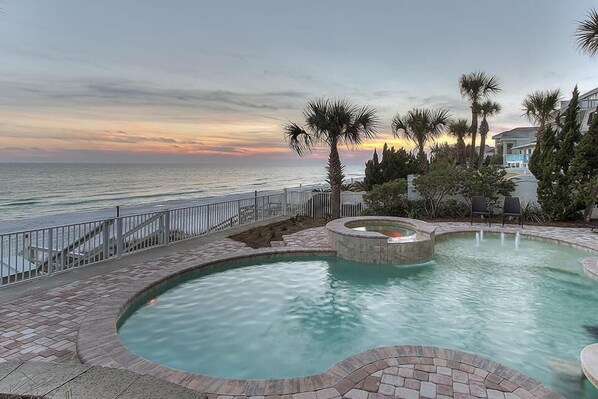 A swimming pool with a view of the ocean at sunset, surrounded by palm trees and a stone deck.