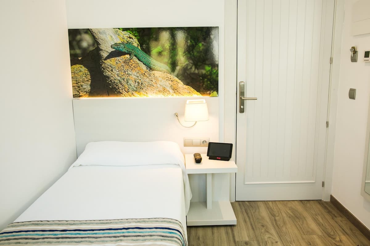 A hotel room with a bed, a nightstand with a lamp, a digital clock, and a painting of a peacock on the wall.