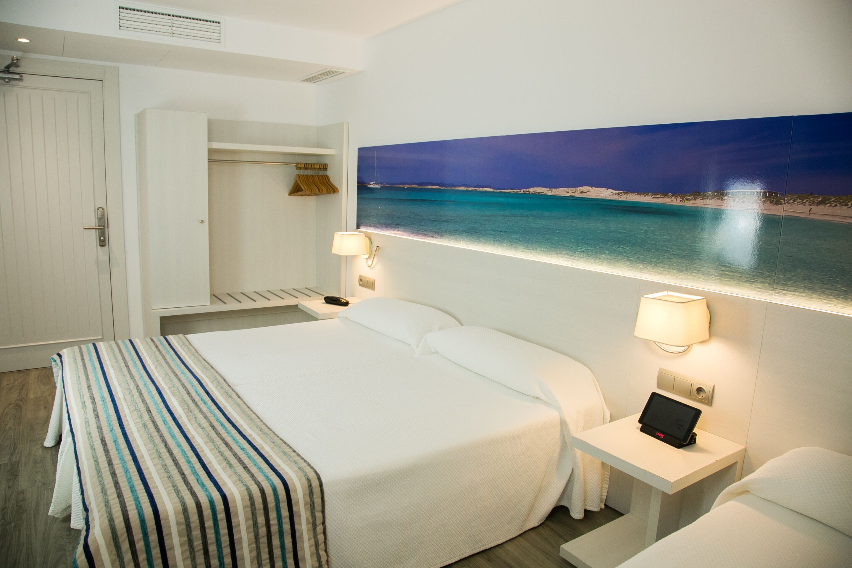 Una camera d'albergo con un letto, comodini, un armadio e un quadro raffigurante una spiaggia.