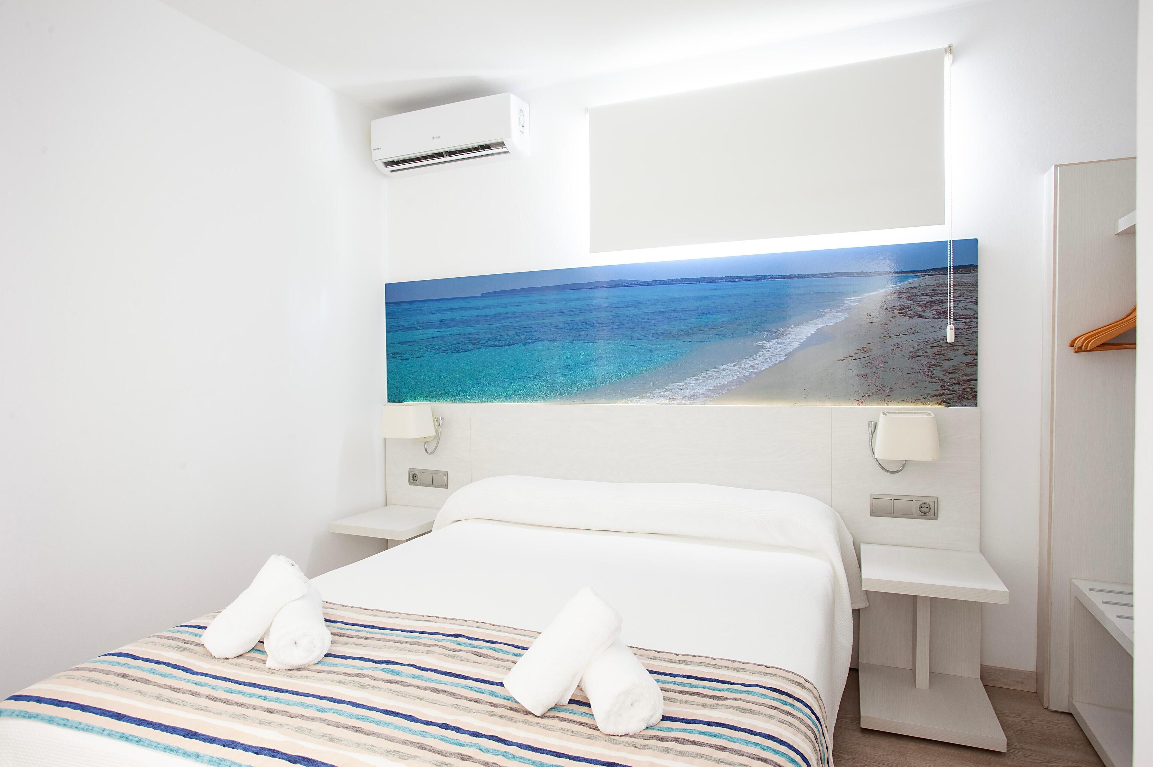 A hotel room with a bed, a large window with a beach view, a bedside table, and an air conditioning unit.
