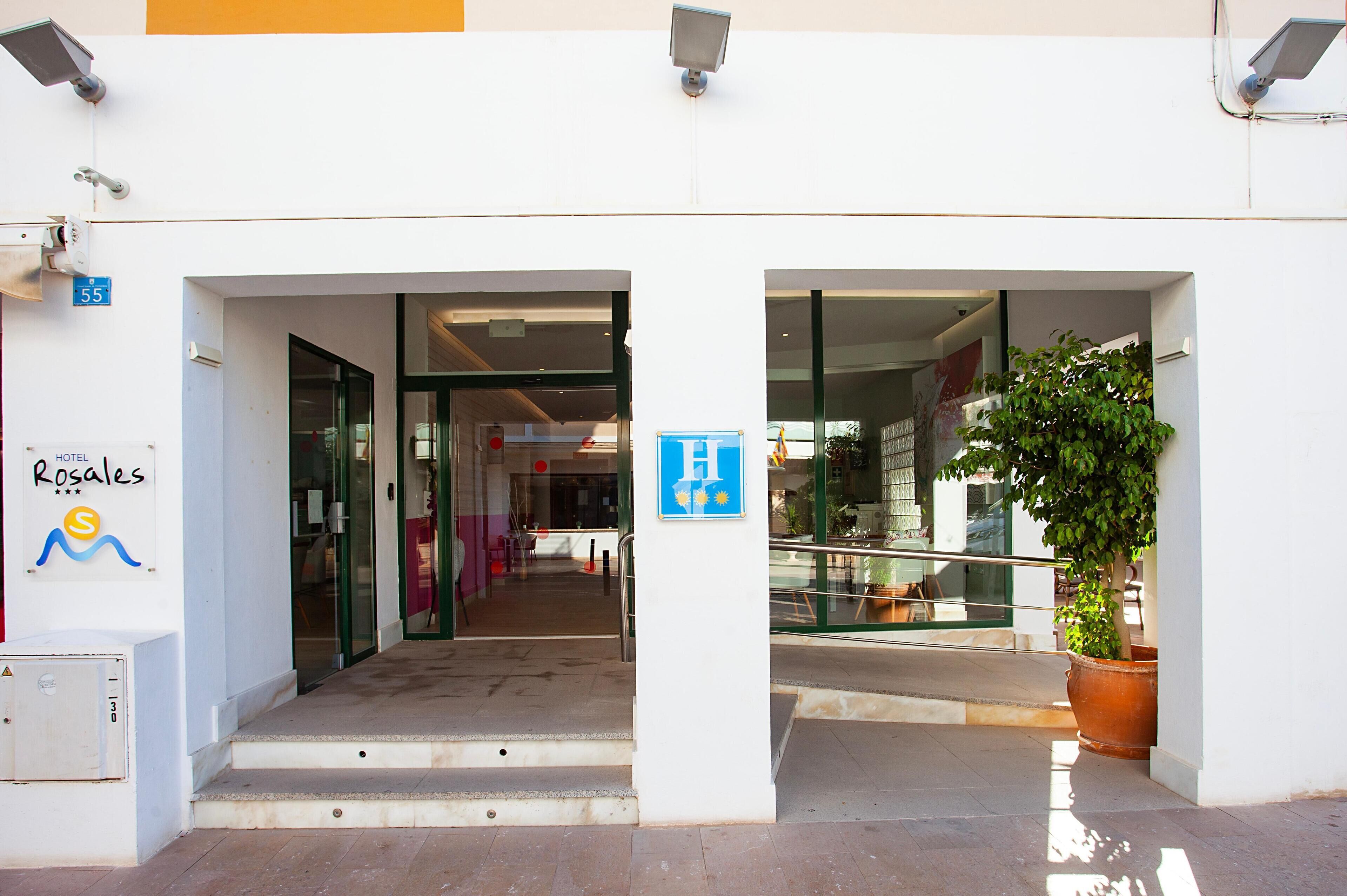 Entrada de hotel con una rampa que lleva a la puerta, un letrero de accesibilidad para personas con discapacidad en azul y una planta en maceta a la derecha.