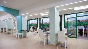 A modern indoor dining area with white chairs and tables, large windows with green frames, and a view of outdoor string lights.