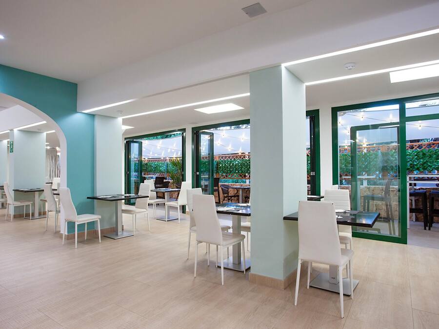 A modern indoor dining area with white chairs and tables, large windows with green frames, and a view of outdoor string lights.