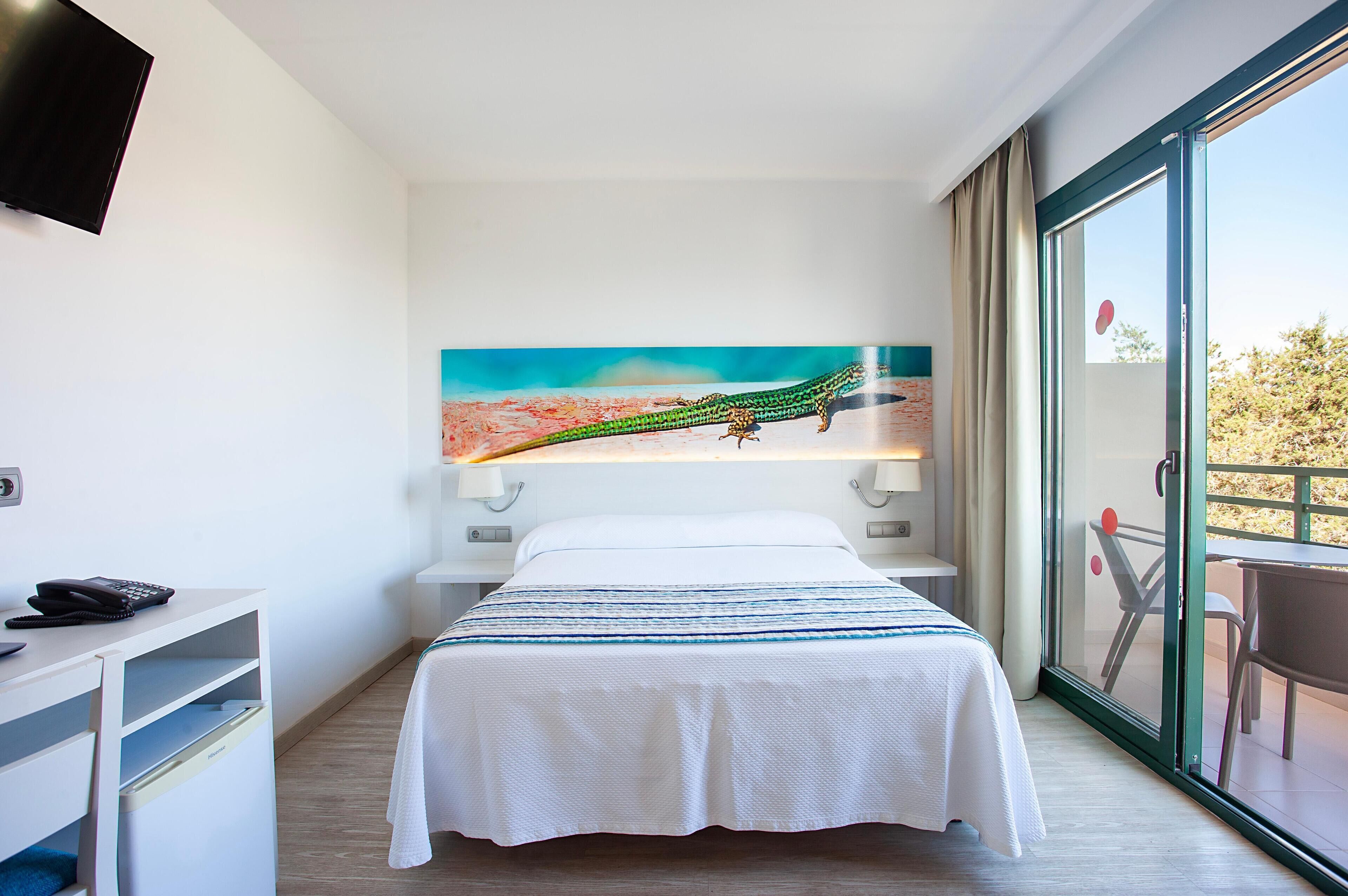 Quarto de hotel com uma cama, uma mesa com telefone, um quadro na parede e uma varanda com mesa e cadeiras.